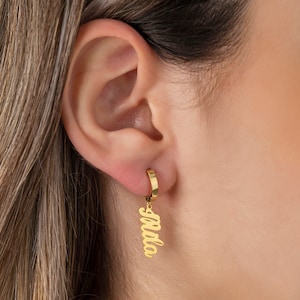 Può includere: Un orecchino a cerchio color oro con un ciondolo pendente. Il ciondolo presenta il nome "Mila" in un carattere corsivo. L'orecchino è indossato sull'orecchio di una persona, con l'attenzione focalizzata sul gioiello.