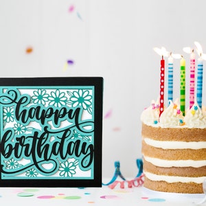May include: A black card with a teal "happy birthday" cutout design with white flowers. A white cake with vanilla frosting and colourful candles is in the background.