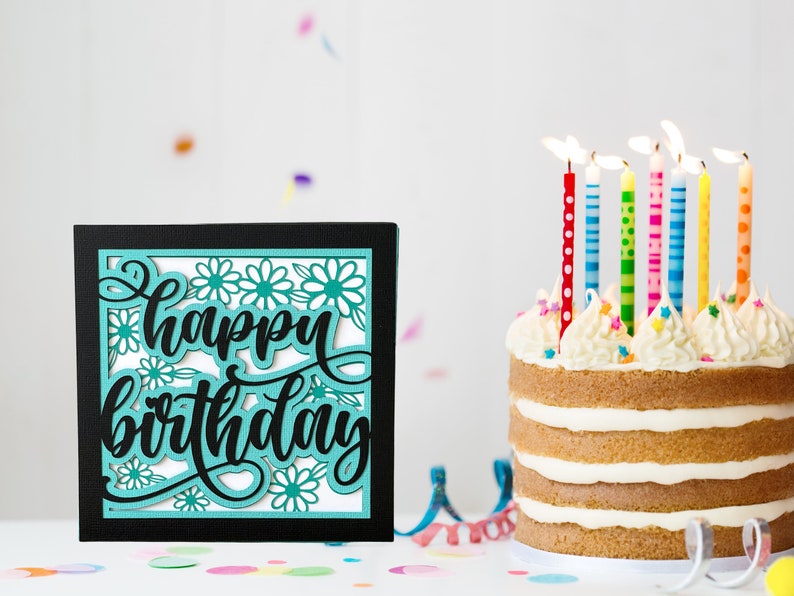 May include: A black card with a teal "happy birthday" cutout design with white flowers. A white cake with vanilla frosting and colourful candles is in the background.