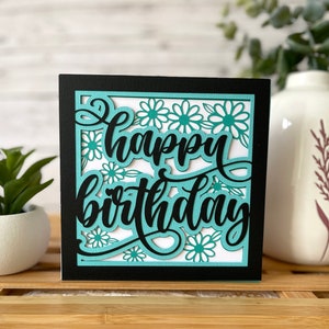 May include: A black card with a white background and a teal cutout design. The card says "happy birthday" in a cursive font.