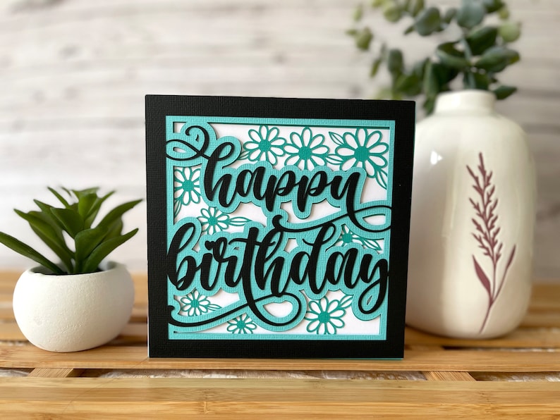 May include: A black card with a white background and a teal cutout design. The card says "happy birthday" in a cursive font.