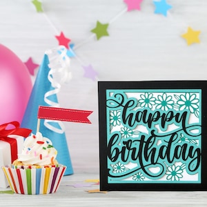 May include: A black card with a turquoise and white cutout design that says "Happy Birthday". The card is sitting on a white wooden surface with a cupcake, a gift box, and a party hat.