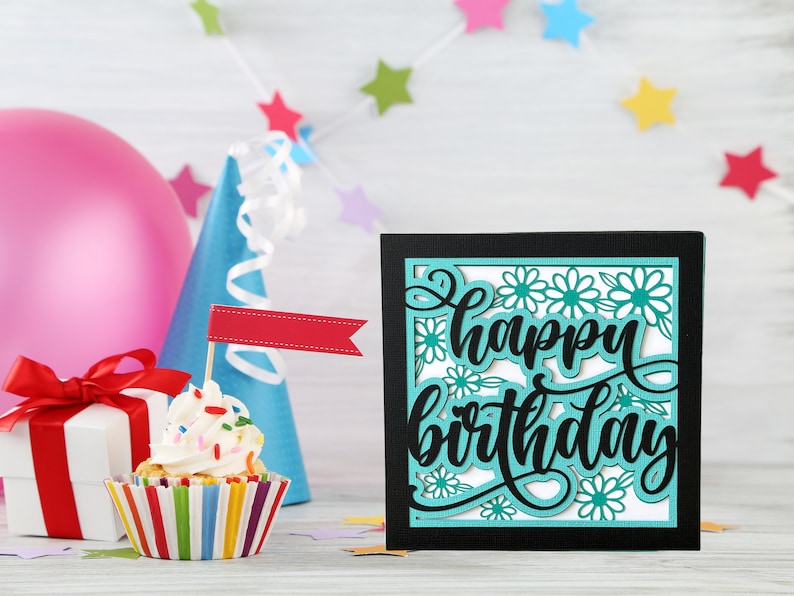 May include: A black card with a turquoise and white cutout design that says "Happy Birthday". The card is sitting on a white wooden surface with a cupcake, a gift box, and a party hat.