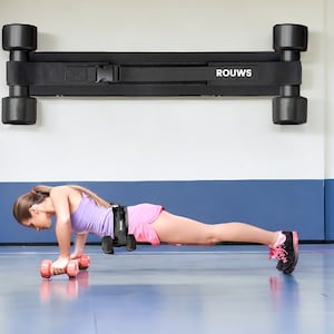 May include: A woman is doing a push-up with a pair of pink dumbbells on a blue and white floor. A black wall-mounted exercise bar with the word "ROUWS" printed on it is in the background.