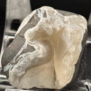 May include: A raw mineral specimen featuring a dark gray, smooth outer rock surface with a contrasting white, crystalline interior cavity. The stone displays natural textures, with translucent white quartz-like formations filling the center.