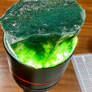 May include: A rough, translucent, emerald-green gemstone is illuminated by a flashlight. The stone has a textured surface and is partially inside the flashlight's opening, revealing a vibrant, lighter green interior. The flashlight is black with a red accent.