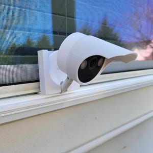 Op de afbeelding: Een witte beveiligingscamera gemonteerd op een raamkozijn. De camera heeft een zwarte lens en een beschermkap. Het raam heeft een scherm en weerspiegelt de lucht en de bomen. De camera is bevestigd aan een witte montagebeugel.