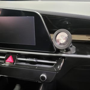 Puede incluir: Primer plano del salpicadero de un coche con una pantalla de infoentretenimiento negra y un objeto circular plateado montado en el salpicadero. El botón de las luces de emergencia es rojo. El salpicadero es negro.