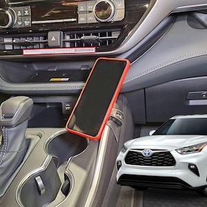 May include: Interior view of a car with a red-framed smartphone mounted on the dashboard. The car's console features a gear shift, cup holders, and a storage compartment. A white Toyota Highlander SUV is visible in the background.