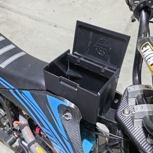 May include: Black storage box with an open lid, mounted on a motorcycle. The motorcycle seat is black with white stripes and blue accents. The handlebars have a yellow and black "PIT BIKE" label.