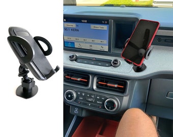 Puede incluir: Un soporte de teléfono negro con una base de ventosa está montado en el tablero de un coche. El soporte de teléfono está sujetando un teléfono inteligente negro. La radio del coche muestra la hora 10:52 y la estación 90.1 KERA.