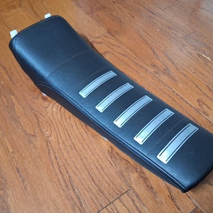 May include: A black motorcycle seat with five silver stripes. The seat has a textured surface and is rectangular. The seat is designed for a motorcycle.