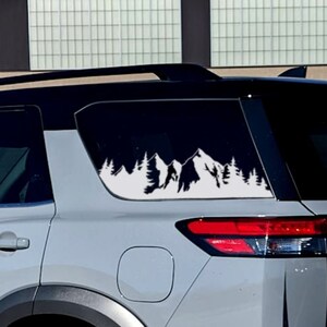 May include: A white vinyl decal of a mountain range with a silhouette of a person fishing in a lake. The decal is applied to the rear window of a grey SUV.