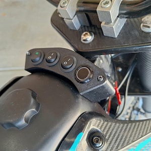 May include: Close-up of a black control panel with three toggle switches and a round button, mounted on a black motorcycle component. The panel is attached to the handlebars with silver hardware. The motorcycle has a carbon fibre design.