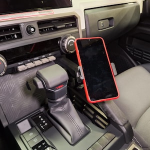 May include: A red-framed smartphone is mounted in a car's interior, next to the gear shift. The car's dashboard features climate controls and a center console with a TRD logo. The interior is grey and black.