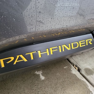 May include: A close-up of a gray car with the word "PATHFINDER" in yellow lettering on the side.