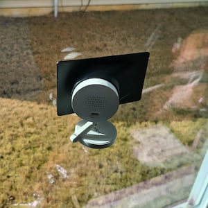 May include: A white and black smart home device with a circular base and a rectangular top, mounted on a window. The device has a speaker and a USB port. The background shows a grassy area.
