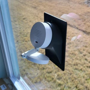 May include: A white and gray security camera mounted on a window. The camera has a circular design with a speaker and a USB port. It is attached to a black rectangular panel and a white adjustable bracket.