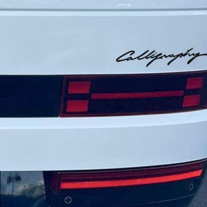 May include: White vehicle rear with black accents and red taillights. The word "Calligraphy" is scripted in black on the vehicle's surface. The image showcases a close-up of the car's design, highlighting the text and the taillight details.
