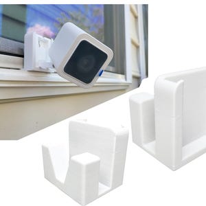 Puede incluir: Juego de cámara de seguridad y soporte de montaje blancos. La cámara es cuadrada con una lente oscura y un acento azul. El soporte está diseñado para montar la cámara en un marco de ventana. También se muestran ejemplos de soportes adicionales.