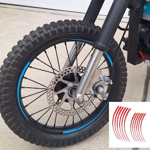 May include: Close-up of a black motorcycle tire with a blue rim stripe. The tire has a knobby tread pattern and is mounted on a black wheel with silver spokes and a disc brake. Red curved decals are in the lower right corner.
