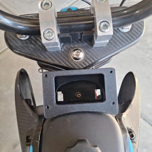May include: Close-up view of a black and silver scooter handlebar assembly. The handlebars are black, with silver mounting brackets. The body of the scooter has a carbon fiber pattern. A black rectangular component is mounted in the center.