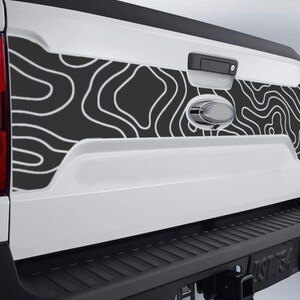 May include: White truck tailgate with a black and white topographic map design. The design is applied to the center panel of the tailgate. The truck has a black bumper and a chrome handle and emblem.