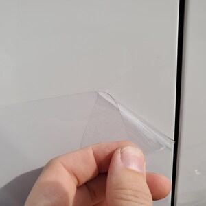 May include: A hand peeling back a clear protective film from a white surface. The film is partially lifted, revealing the surface underneath. The image is well-lit, showing the texture of the film and the white panel.