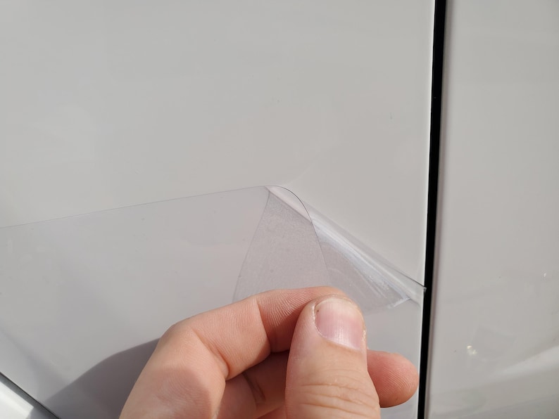 May include: A hand peeling back a clear protective film from a white surface. The film is partially lifted, revealing the surface underneath. The image is well-lit, showing the texture of the film and the white panel.
