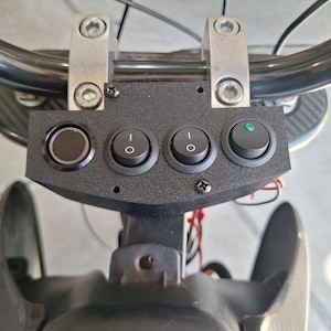 May include: Close-up of a motorcycle handlebar with a black control panel. The panel has four switches, including a black button and three toggle switches. The handlebar is black, and the mounting brackets are silver.