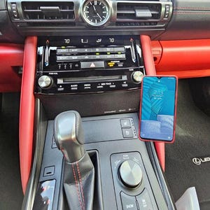 Puede incluir: Interior de coche con un smartphone montado en el salpicadero. El salpicadero incluye un reloj, radio y controles de climatización. La pantalla del teléfono muestra una imagen azul y blanca. Se ven detalles en rojo.