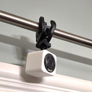 May include: A small, white square camera is mounted on a black clamp, attached to a silver metal rod. The camera lens is visible, and the setup is against a light gray wall and white trim.