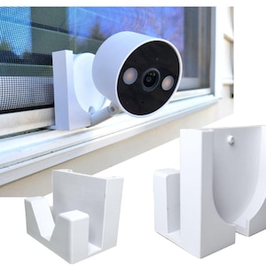 Puede incluir: Una cámara de seguridad blanca montada en una ventana, con una lente negra y dos lentes más pequeñas. También se muestran dos soportes de montaje blancos. La cámara está diseñada para la seguridad y vigilancia del hogar.