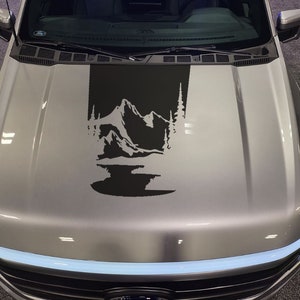 Può includere: Un pick-up argentato con un adesivo nero di una scena di montagna sul cofano. L'adesivo presenta una catena montuosa, alberi e un lago.