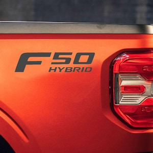 May include: Close-up of the rear of an orange truck with the text "F50 HYBRID" in black. The truck's taillight is visible on the right side, featuring red and white lights. The vehicle's design is modern and sleek.