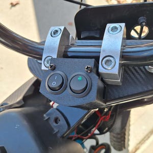 May include: A close-up of a black handlebar-mounted control panel with two round switches. One switch is labeled with a circle and a line, the other has a green light illuminated. The panel is mounted with silver brackets.
