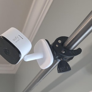 May include: A white and black security camera with the brand name "eufy" is mounted on a metal pole using a black clamp. The camera is attached to a white base with a cylindrical connector.