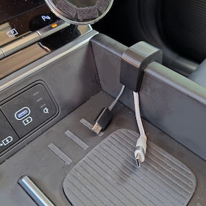 May include: A black, 3D-printed car phone charger holder with a USB-C cable attached. The holder is clipped onto the edge of a car console. The console has buttons and a wireless charging pad.