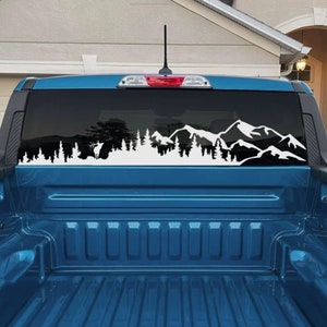 May include: A blue pickup truck bed with a white vinyl decal of a mountain range and trees on the rear window. The decal features a silhouette of a deer in the forest.