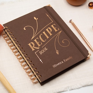Può includere: Un ricettario marrone rilegato a spirale con scritte dorate che recitano "RECIPE BOOK" e "Oliveira Family". Una penna dorata è appoggiata accanto al libro. Il libro è su una superficie bianca con un cucchiaio di legno e un panno arancione.