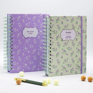 Puede incluir: Dos agendas de espiral con estampados florales. La agenda de la izquierda es morada con el texto PLANNER 2027. La agenda de la derecha es verde claro con el texto MARIE PLANNER 2026-2027 y tiene una banda elástica de cierre morada en el lateral.