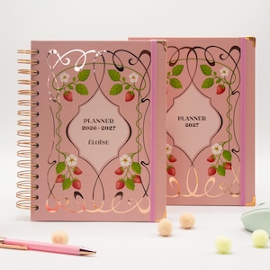 Op de afbeelding: Twee roze planners met spiraalbinding, voorzien van een decoratief kader met aardbei-illustraties en witte bloemen. Op de voorkant staat de tekst PLANNER 2026-2027 ÉLOÏSE en PLANNER 2027. De planners hebben een roségouden spiraal en roze elastieken.