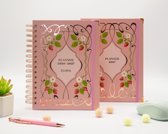 May include: Two pink spiral-bound planners featuring a decorative frame with strawberry illustrations and white flowers. The front cover displays the text PLANNER 2026-2027 ÉLOÏSE and PLANNER 2027. The planners have rose gold wire binding and pink elastic bands.