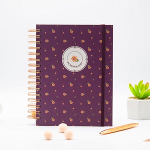 Puede incluir: Una agenda de espiral de color morado oscuro con un patrón repetitivo de pequeñas flores naranjas. La portada presenta una etiqueta circular central con una ilustración floral y el texto PLANNER 2027. Incluye una espiral de color cobre y una banda elástica.