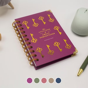 Op de afbeelding: Een magenta spiraalgebonden wachtwoordenboek met de titel The Little Book of Great Forgettings, voorzien van een gouden sleutelpatroon, gouden hoekbeschermers en een gouden spiraal. De tekst staat in gouden letters op de magenta kaft.