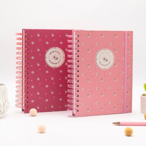 Op de afbeelding: Twee spiraalgebonden planners met een bloemenpatroon. De linker planner is donkerroze en de rechter is lichtroze met een verticaal elastiek. Beide hebben een centraal rond label met de tekst "YOUR NAME PLANNER 2021" of "PLANNER 2021".