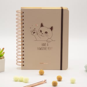 Peut inclure: Un agenda beige à reliure spirale avec une spirale en cuivre et un élastique de fermeture marron foncé. La couverture présente une illustration au trait d'un chat qui fait coucou et le texte "HAVE A PAWSITIVE YEAR!" et "PLANNER 2021" en cuivre.