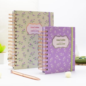 May include: Two floral notebooks with spiral bindings. One is light green, the other purple. Both feature the name "Ana Luisa" and "AGENDA 2020". A rose gold pen is placed nearby. The notebooks are likely for planning.
