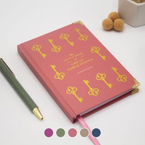 Puede incluir: Un libro de contraseñas rectangular con una cubierta de color rosa empolvado que presenta un patrón repetitivo de llaves doradas. El texto en la cubierta dice The Little Book of Great Forgettings. Incluye esquinas metálicas doradas y un marcapáginas.