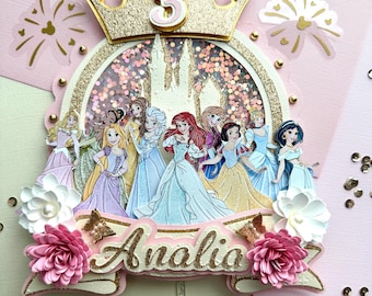May include: A pink and gold cake topper featuring a princess castle with a glitter background, a crown with the number 3, and a banner with the name "Aalia".